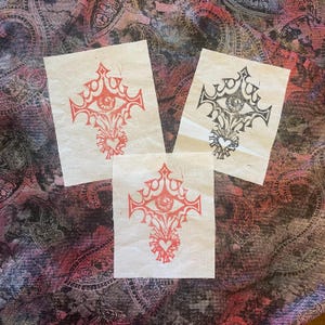 May include: Three square prints with a cross design, an eye at the centre, and a heart below. Two are red, one is black, all on off-white paper. The background is a paisley pattern in shades of red, pink, and black.