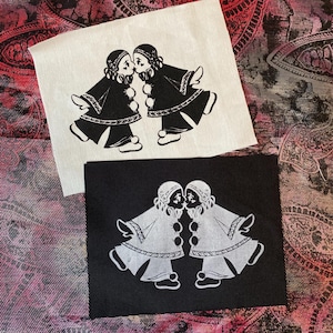 Puede incluir: Dos parches con una ilustración en blanco y negro de dos figuras besándose. Un parche es blanco con un diseño negro, y el otro es negro con un diseño blanco. Las figuras llevan sombreros y abrigos.
