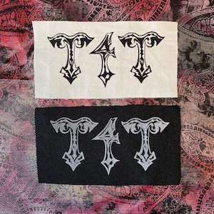 May include: Two rectangular patches with the letters "T4T" in a gothic font. One patch is white with black lettering, and the other is black with silver lettering. The patches are likely for clothing or accessories.