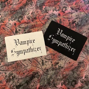 May include: Two rectangular patches with the text "Vampire Sympathizer" in a gothic font. One patch is black with white text, and the other is off-white with black text. The background is a patterned fabric with red, pink, and gray paisley designs.