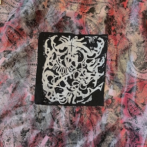 Könnte beinhalten: Ein schwarzer quadratischer Aufnäher mit einem silbernen, abstrakten Design mit einem Totenkopf und einem Kreuz. Der Aufnäher befindet sich auf einem farbenfrohen Paisley-Stoffhintergrund mit Rosa-, Rot- und Grautönen.