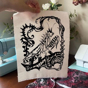 Peut inclure: Une pièce rectangulaire de tissu en toile blanc cassé présentant une illustration sérigraphiée en noir d'une mante religieuse portant des bottes à lacets. L'insecte est encadré par des vignes épineuses stylisées sur une forme organique.