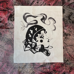 May include: A black and white fabric patch featuring a vintage-style illustration of a woman with a headscarf, smoking. The design includes a stylized smoke cloud and a decorative collar. The patch is on a white background.