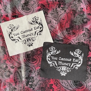 Pode incluir: Duas pequenas bolsas quadradas, uma creme e outra preta, com o texto "You Cannot Eat Money" em uma fonte decorativa. O texto é emoldurado por desenhos florais e de rolagem. O fundo é um padrão paisley em tons de rosa, vermelho e cinza.