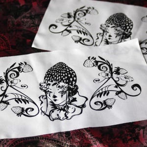 以下が含まれることがあります： イチゴ柄の帽子をかぶった中央の人物と、その左右に描かれたイチゴの蔓、葉、花の黒いインクイラストが施された、2枚の長方形の白い布製パッチ。顔の両側に装飾的な植物のモチーフが配置されている。
