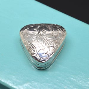 May include: A small, triangular, silver-coloured metal trinket box featuring an intricate, engraved floral scrollwork pattern on its lid. The surface of the box is polished and reflective, showcasing detailed etched lines and a decorative beaded border.