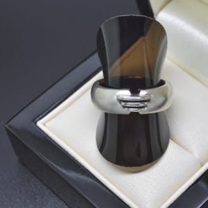 May include: A silver ring with a unique design, displayed in a black jewelry box with a white interior. The ring features a wide band with a layered, geometric detail in the center. The ring is showcased on a dark brown stand.