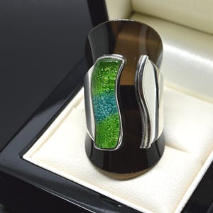 May include: A wide silver ring featuring a curved, vertical inlay of textured glass in shades of lime green and teal. The glass surface displays a bubbly, organic pattern, contrasted by the smooth, polished silver band that wraps around the finger.