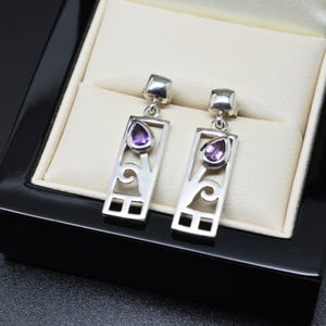 May include: A pair of silver drop earrings featuring a rectangular frame with an openwork scroll design and a single teardrop-shaped purple amethyst gemstone set near the top of each piece, suspended from a small square silver post attachment.