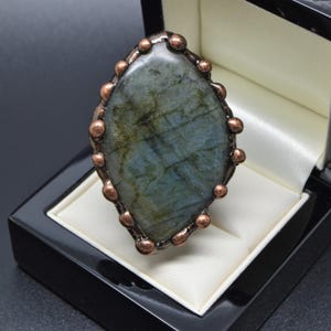 May include: An oval ring with a large labradorite stone, displaying iridescent colours. The stone is set in a copper-toned metal frame with decorative details. The ring is presented in a black jewellery box with a white satin lining.