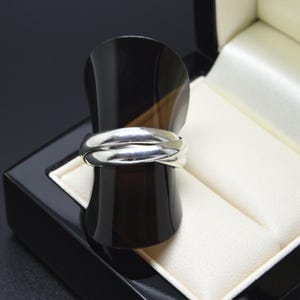 May include: A silver ring featuring three interlocking bands with a polished, reflective finish. The ring is displayed on a curved, dark brown stand that is positioned inside a black jewellery box with a cream-coloured interior lining.