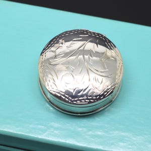May include: A small, round, silver-coloured metal pill box featuring an intricate, engraved floral pattern on its lid. The surface of the lid is polished and reflective, showcasing detailed scrollwork and leaf motifs across the top circular face.