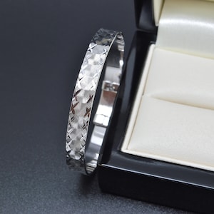 May include: A silver bangle bracelet with a textured, geometric pattern. The bracelet is displayed next to a black jewelry box with a white satin interior. The bangle is a classic piece of jewelry.