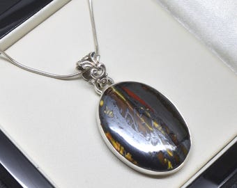 May include: An oval-shaped pendant featuring a polished, dark stone with streaks of gold, brown, and red, set in a silver frame. The pendant hangs from a thin silver snake chain, with an ornate, decorative silver bail connecting the two pieces.