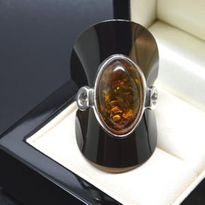 May include: A silver ring featuring a large, oval-shaped, translucent amber gemstone with deep golden and brown inclusions. The stone is set in a smooth silver bezel with decorative circular accents on each side of the band, displayed upright.