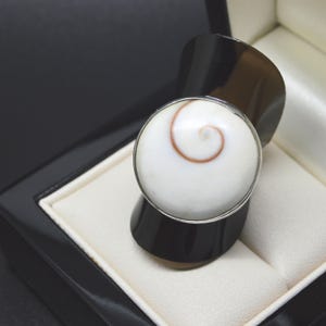 Op de afbeelding: Een ring met een ronde witte shiva-oogschelp met een natuurlijk bruin spiraalpatroon, gezet in een zilverkleurige metalen band. De band heeft een brede, gladde, donker-metalen afwerking die naar boven buigt om het ronde middelpunt te dragen.