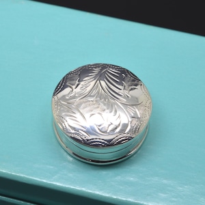 May include: A small, round, silver-coloured metal pill box featuring an intricate, engraved floral pattern on its lid. The surface of the lid is highly reflective and polished, showcasing detailed scrollwork and cross-hatched textures.