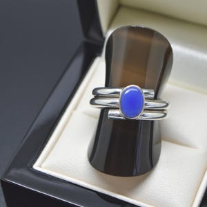May include: A silver ring featuring a smooth, polished band and a central, oval-shaped blue cabochon stone. The ring has a sleek, metallic finish and a minimalist design, showcasing a vibrant blue gemstone set securely within the silver metal frame.