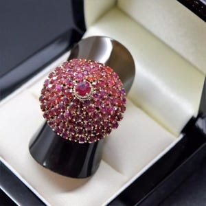 May include: A gold ring featuring a large, round ruby gemstone encircled by smaller rubies. The ring is set in a white metal band and is presented in a black ring box.