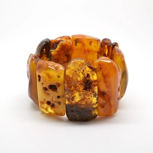 May include: A stretch bracelet composed of multiple polished, irregular rectangular pieces of amber. The segments display a range of warm, translucent honey, golden, and deep brown tones, featuring natural inclusions and varied surface textures.