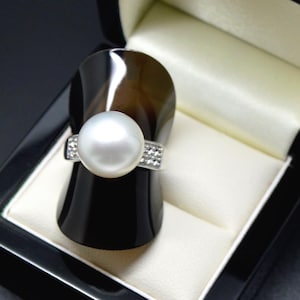 Op de afbeelding: Een zilveren ring met een grote, ronde, witte parel in het midden van de band. De band is versierd met kleine, heldere, fonkelende steentjes die aan de zijkanten van de zetting zijn ingelegd, wat een helder contrast vormt met de gladde parel.