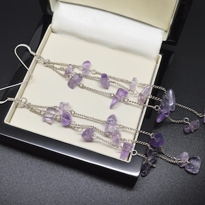 May include: A pair of long dangle earrings featuring multiple silver-toned chains adorned with small, irregular, translucent purple amethyst gemstone chips. The earrings are attached to silver-toned ear wires and are displayed in a black jewelry box.
