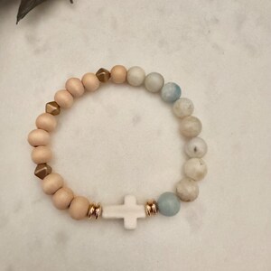 May include: A bracelet with a white cross charm and a mix of light pink, gold, and blue-green beads. The bracelet is made with wooden beads and gold-coloured spacers. The cross is centered on the bracelet.