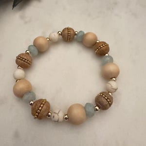 Peut inclure: Un bracelet de perles composé de perles en bois naturel, blanches et bleu clair. Le bracelet comprend des perles rondes en bois brun clair, des perles marron texturées et de petites perles dorées. Le bracelet est sur une surface blanche.