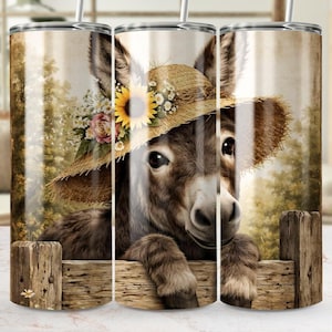 May include: A tumbler featuring a charming donkey wearing a straw hat adorned with a sunflower and floral accents. The donkey is depicted behind a wooden fence, set against a soft, blurred background. The tumbler is designed for beverages.