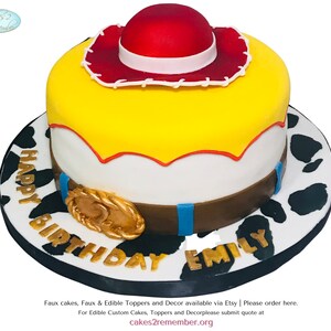 May include: A yellow and white cake decorated to look like Woody from the Toy Story movie. The cake has a red cowboy hat, a brown belt with a gold buckle, and a white and black cow print base. The cake is inscribed with "HAPPY BIRTHDAY EMILY".