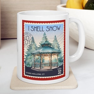 May include: White ceramic mug with a red and white postage stamp design. The stamp features a snowy scene with a gazebo and the text "I Smell Snow" and "Stars Hollow, CT".
