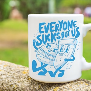 May include: White ceramic mug with a blue graphic of a burger and a slice of pizza with the text "Everyone Sucks But Us".