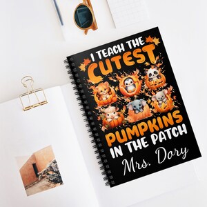 Könnte beinhalten: Ein schwarz gebundenes Notizbuch mit dem Text "I Teach The Cutest Pumpkins In The Patch Mrs. Dory" auf dem Cover. Das Cover zeigt Illustrationen von Tieren in Kürbissen. Das Notizbuch liegt offen auf einer weißen Oberfläche.