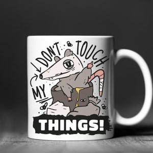 May include: White ceramic mug featuring a cartoon opossum inside a tree stump, giving the middle finger. The mug has the text "I DON'T TOUCH MY THINGS!" in black and white, with additional cartoon elements.
