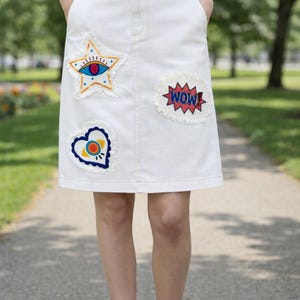 May include: White denim skirt with colorful embroidered patches. The patches include a star with an eye, a heart, and a "WOW" speech bubble. The skirt has a classic five-pocket design and is worn with a denim shirt.