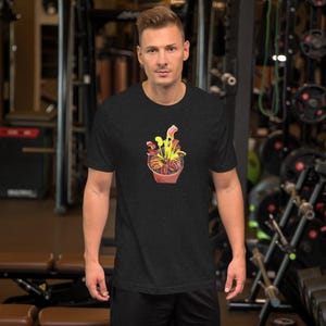 May include: Dark gray t-shirt featuring a colorful illustration of a potted plant. The plant has yellow, red, and green leaves and is in a terracotta-colored pot. The shirt is short-sleeved and has a crew neck.