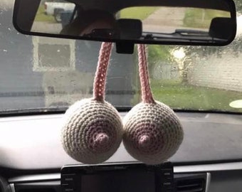 May include: A pair of crocheted pink and white breast-shaped car air fresheners hanging from the rearview mirror of a car.
