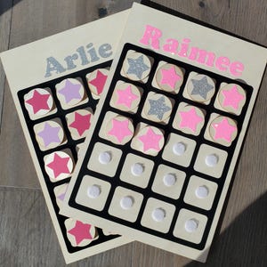 May include: Two wooden boards with black grid patterns. Each board has a name in glitter letters: "Arlie" and "Raimee". The grids contain star-shaped and circular pieces in pink, purple, and silver glitter. White circular pieces are also present.