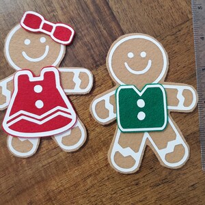 May include: Two gingerbread man ornaments. One has a red bow and dress, the other a green vest. Both have white icing details and smiling faces. The ornaments are made of felt and are on a wooden surface.