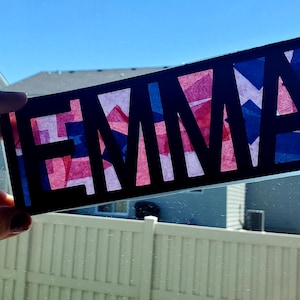 May include: A black rectangular sign with the name "EMMA" cut out, revealing red, white, and blue tissue paper. The sign is held up against a bright blue sky, with a white fence and houses in the background.