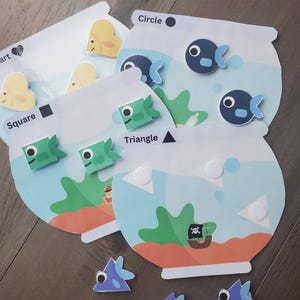 May include: Educational toy set featuring fish-shaped magnets and cards with shapes and the words "Circle", "Square", "Triangle", and "Heart". The fish magnets are blue, green, and yellow. The cards have a fishbowl design.
