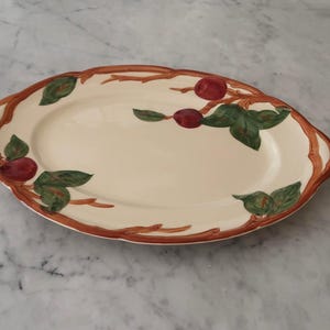 May include: An oval-shaped cream-colored ceramic serving platter featuring a hand-painted border of brown tree branches, green leaves, and small red fruit. The platter has integrated handles on both sides and a smooth, glossy finish.