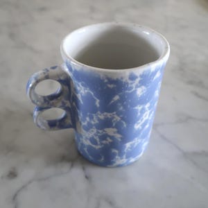 May include: A small, blue and white speckled ceramic mug with a unique double-loop handle. The mug has a cylindrical shape and a white interior. The exterior features a light blue base with white speckles, creating a marbled effect. This mug is perfect for coffee or tea.