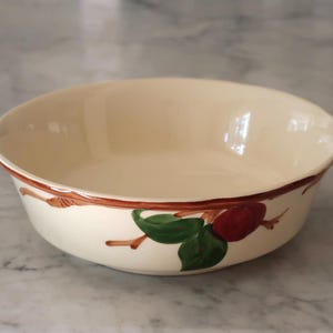 May include: A round, cream-colored ceramic bowl featuring a hand-painted design around the exterior rim. The pattern consists of a thin brown branch with vibrant green leaves and a single dark red fruit, creating a simple, decorative border.