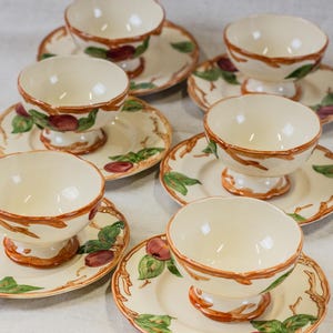 May include: A set of cream-colored ceramic dessert bowls and saucers. Each bowl has a pedestal base and is encircled with a brown rim. The saucers and bowls are decorated with a fruit and leaf design in red, green, and brown.
