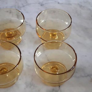 May include: Four amber-colored, stemmed glasses with rounded bowls. The glasses have a warm, golden hue and sit on a marble surface. The stem and base are a darker shade of amber, creating a contrast.