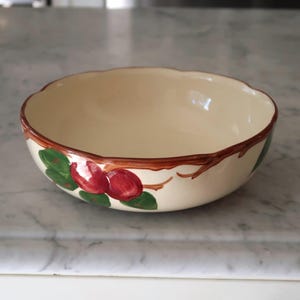May include: A round ceramic bowl with a cream-colored interior and exterior. The exterior features a hand-painted design of brown tree branches with green leaves and red apples encircling the rim. The bowl has a scalloped edge along the top.