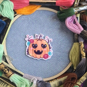 Puede incluir: Un bastidor de punto de cruz azul claro con una pegatina de calabaza en el centro, rodeado de hilo de bordar colorido. La pegatina de calabaza es naranja con ojos en forma de corazón y detalles florales. Los colores del hilo incluyen rosa, verde, amarillo y azul.