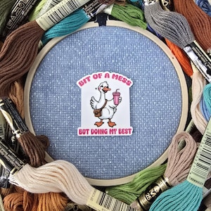 May include: A cross-stitch hoop with a light blue fabric, surrounded by colorful embroidery floss. A sticker in the center reads "Bit of a Mess But Doing My Best," featuring a cartoon goose. The floss colors include orange, blue, green, and brown.