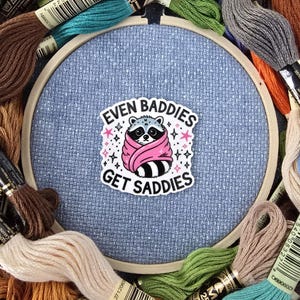 May include: A cross-stitch hoop with a light blue fabric, surrounded by colorful embroidery floss. The hoop features a sticker with a raccoon wrapped in a pink scarf and the text "EVEN BADDIES GET SADDIES".